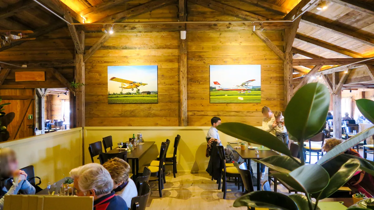 Die Fliegerthematik will man im neuen Restaurant gezielt integrieren. Man sieht das Innere eines Holzchalets mit Gästen.